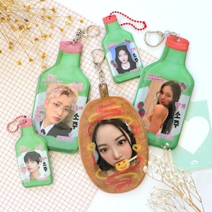 May include: Five colorful keychains featuring different images of people. The keychains are shaped like green bottles with pink accents and the text "소주" which means "soju" in Korean. One keychain is shaped like a brown oval with a picture of a person eating a hot dog.