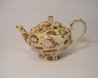 May include: A yellow ceramic teapot with a lid, spout, and handle. The teapot is decorated with Christmas-themed illustrations, including wreaths, birds, and bows. The handle and spout are trimmed with gold.