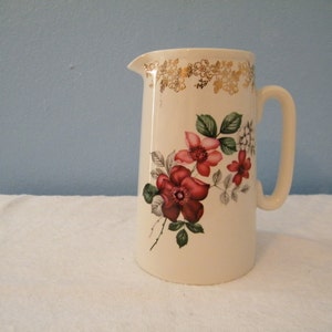 May include: A white ceramic pitcher with a gold trim and a floral design featuring red, pink, and green flowers.
