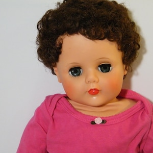 May include: A vintage doll with brown curly hair, green eyes, and rosy cheeks. The doll is wearing a pink long-sleeved shirt with a small flower detail at the neckline. The doll's lips are painted red.
