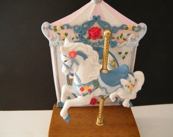 May include: A white carousel horse with a blue saddle and floral details, mounted on a gold pole. The horse is part of a miniature carousel with a pink and white canopy.