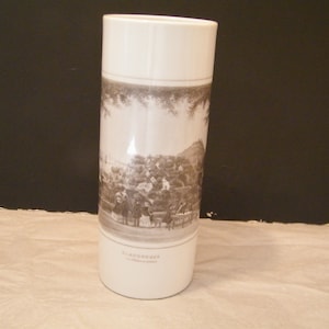 May include: White ceramic vase with a cylindrical form. The vase has a black and white illustration of a historical scene. The top and bottom sections are plain white. The vase has text at the bottom.