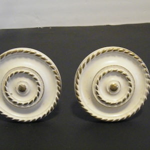 May include: Two off-white cabinet knobs with a gold rope-like trim around the edge and concentric circles. The center of each knob has a small, gold-colored accent. These decorative knobs are likely for furniture.