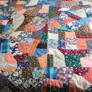 May include: A patchwork quilt with a variety of fabric patterns and colors. The quilt features squares and irregular shapes in shades of blue, red, green, and floral prints. The quilt is a handmade item.