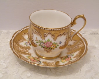 May include: A white teacup and saucer with gold trim and floral designs. The cup has a delicate handle and the saucer has a scalloped edge.