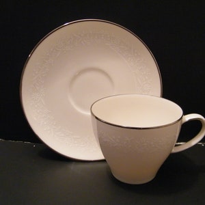 May include: A white teacup and saucer set with a floral pattern and a gold rim. The teacup has a curved handle and sits next to its matching saucer. The set is on a dark surface.
