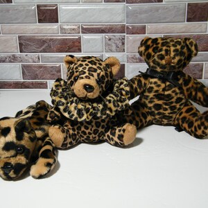 May include: Three plush leopard-print teddy bears. One bear is lying down, another is sitting with a decorative collar, and the third is sitting upright with a black bow. The bears have tan and black spots.