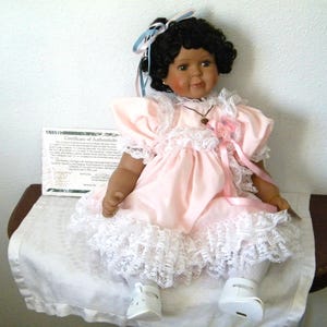 May include: A porcelain doll with dark curly hair, wearing a light pink dress with white lace trim and a matching ribbon in the hair. The doll features a small necklace and white shoes. A Certificate of Authenticity document is positioned behind the doll.