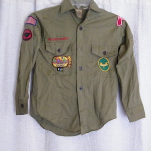 May include: Olive green long-sleeve shirt with multiple patches. Patches include an American flag, a red and black emblem, and a Tumwater Council patch. The shirt has two chest pockets and buttons down the front. The words "Boy Scouts of America" are embroidered on the shirt.