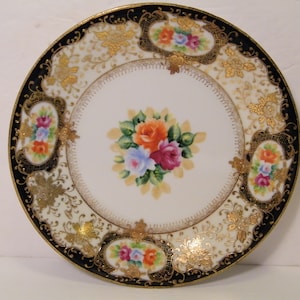 May include: A white porcelain plate with a black and gold rim. The center of the plate features a floral design with pink, orange, and blue flowers. The rim is decorated with a gold floral pattern.