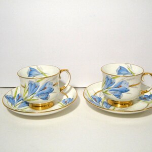 May include: Two matching teacups and saucers. The white porcelain cups and saucers are decorated with blue floral designs and gold trim. The cups have gold handles and sit on gold bases. The saucers are round and have a matching floral pattern.