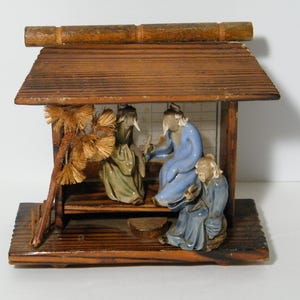 May include: A wooden miniature house with a thatched roof, featuring four ceramic figurines of people sitting inside. The figurines are wearing traditional clothing and have painted details. The house is decorated with a small pine tree.