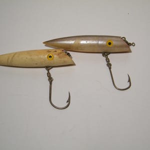 May include: Two wooden fishing lures with yellow eyes and silver hooks. The lures are different colours, one is light brown and the other is silver.