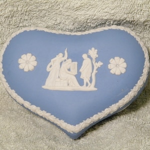 May include: A heart-shaped, light blue trinket box with a white relief design. The box features a white floral border and two white flower accents. The relief depicts figures in classical attire. A decorative item.