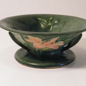 May include: A green ceramic bowl with a pink floral design and two handles. The bowl is on a pedestal base.