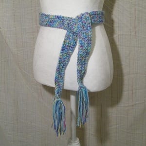 May include: A handmade, multi-colored crochet belt with a tie closure and tassels. The belt is displayed on a white mannequin. The belt's colors include shades of blue, purple, and yellow. The tassels hang down from the belt.