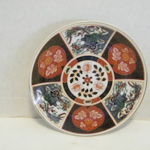 May include: A round, white porcelain plate with a decorative design. The plate features a central floral motif surrounded by six panels, each with a different design. The panels feature a mix of floral, geometric, and landscape patterns in shades of red, black, green, and gold.