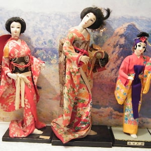 May include: Three traditional Japanese dolls dressed in colorful kimonos. The dolls are standing on wooden bases. The doll on the right is holding a fan and has the text "美人人形" on the base.