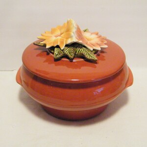 May include: A terracotta-colored ceramic casserole dish with a lid. The lid features a three-dimensional floral design with orange and white flowers and green leaves.