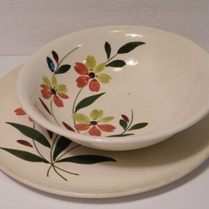 May include: Cream-colored ceramic bowl and plate set. The bowl sits atop the plate. Both pieces are decorated with a floral design featuring orange, yellow, and green flowers with dark green leaves. The set has a vintage aesthetic.