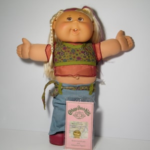 May include: A Cabbage Patch Kids doll with blonde hair and pink highlights wearing a green and pink floral print top, blue jeans, and a pink headband. The doll is holding out both arms and has a big smile on its face. The doll is holding a birth certificate and adoption papers.