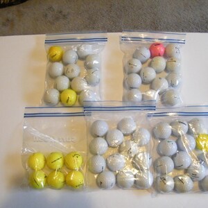 May include: Several clear plastic zip bags filled with golf balls. Most balls are white, with a few yellow and one pink ball visible. The bags are arranged on a white surface, suggesting a sports or recreation theme.