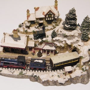 May include: A miniature winter village scene with a train, a house, and snow-covered trees. The train is blue and black with a white 'SDJR' logo. The house is white with a brown roof and a black door. The trees are green and covered in white snow.