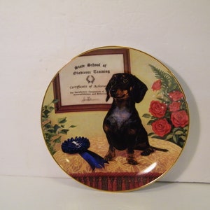May include: A decorative ceramic plate featuring a dachshund dog with a blue ribbon. The plate has a gold rim and a picture of a certificate that reads "State School of Obedience Training, Certificate of Achievement".