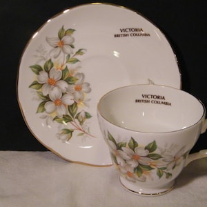 May include: A white teacup and saucer with a gold rim. The teacup and saucer feature a floral design with white flowers and green leaves. The text "VICTORIA BRITISH COLUMBIA" is printed on both the teacup and saucer.
