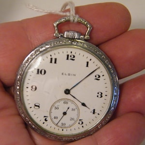 May include: A silver pocket watch with a white face and black Roman numeral hour markers. The watch has a small second hand dial at the bottom of the face. The watch face says "ELGIN".