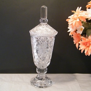 May include: A clear, cut-glass decorative jar with a lid, featuring floral and geometric patterns. The jar stands on a pedestal base and has a tall, rectangular finial. The background is dark, with a bouquet of peach and white flowers.