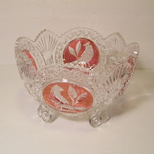May include: Clear glass bowl with a scalloped rim and three small feet. The bowl features etched bird and leaf designs within oval, red-colored panels. The bowl has a cut glass pattern throughout.