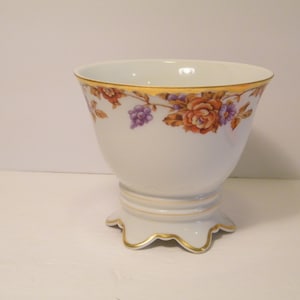 May include: A white porcelain vase with a gold rim and floral design. The vase has a scalloped base and three feet.