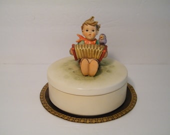 May include: A porcelain figurine of a boy playing an accordion with a bird perched on his shoulder. The figurine is on top of a round, white music box with a brown base.