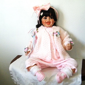 May include: A porcelain doll with black hair, wearing a pink dress, sweater, and pants. The doll has a pink bow, a headband, and pink shoes. The doll is decorated with floral accents and is sitting on a white cloth.
