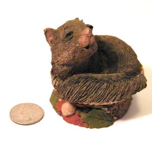 May include: A detailed figurine of a squirrel, primarily brown with a textured fur appearance, curled up on a base with leaves and a small nut. A quarter coin is next to the figurine for size reference.