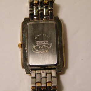 May include: Back of a silver and gold watch with a square face. The watch face has the text "Japan Movt Regent Gol 71 Stainless Steel Back" on it.