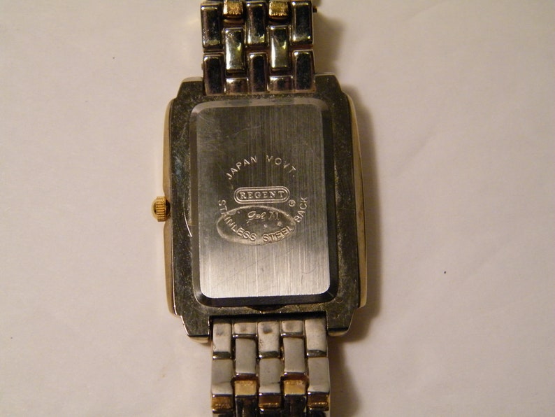May include: Back of a silver and gold watch with a square face. The watch face has the text "Japan Movt Regent Gol 71 Stainless Steel Back" on it.