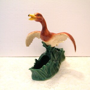 May include: A ceramic planter shaped like a brown and white goose with its wings spread wide, perched atop a dark green base molded to resemble tall grass or reeds. The goose has a yellow beak and is depicted in a dynamic, open-mouthed pose.