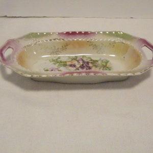 May include: Oval-shaped, antique porcelain dish with a floral design. The dish features a scalloped edge with pink and green accents, gold trim, and two handles. The interior displays a bouquet of purple and yellow flowers.