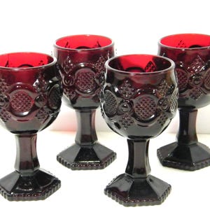 May include: Four ruby red goblets with ornate, textured designs. The glasses have a detailed pattern on the bowl and a faceted base. The interior of the glasses is a vibrant red colour.