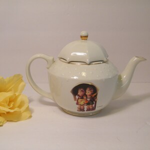May include: Cream-colored porcelain teapot with a lid and a handle. The teapot features a decorative image of two children under an umbrella. The lid is topped with a gold-colored finial. A yellow rose is in the background.