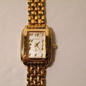 May include: A gold-tone Regent quartz wristwatch with a white face and black Roman numeral hour markers. The watch has a gold-tone metal band.