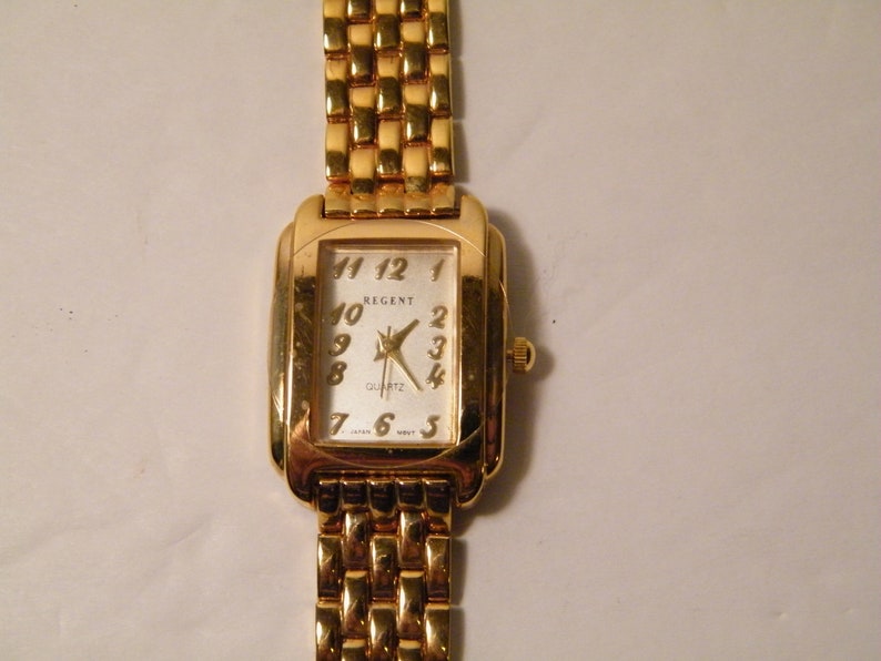 May include: A gold-tone Regent quartz wristwatch with a white face and black Roman numeral hour markers. The watch has a gold-tone metal band.