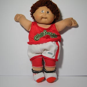 May include: A Cabbage Patch Kids doll wearing a red mesh tank top with the words "Cabbage Patch Kids" in green and white. The doll is also wearing white shorts with red trim, red leg warmers, and white socks with red accents.