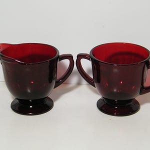 Peut inclure: Un ensemble de deux pots à crème et à sucre en verre rouge avec des anses. Les bols ont un pied.