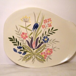 May include: An off-white, asymmetrical ceramic serving platter featuring a central botanical illustration. The design includes stylized flowers in shades of pink, blue, and purple, accented by green leaves and stems, all set against a textured surface.