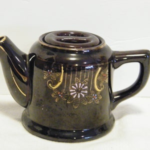 May include: A small, dark brown teapot with a gold floral design and a brown lid. The teapot has a handle and a spout.
