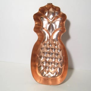 May include: A vintage copper pineapple-shaped mold. The mold has a detailed design with a textured surface, showcasing the pineapple's form. The copper has a warm, reflective hue, adding to its antique charm. A small loop is at the top.
