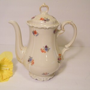May include: Cream-colored porcelain coffee pot with a lid, handle, and spout. The pot is decorated with small floral clusters in orange, purple, and blue. A yellow daffodil is in the lower left corner.